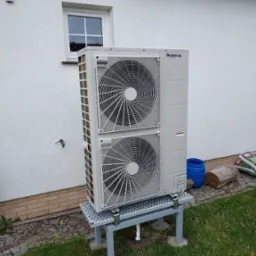 Zewnętrzna jednostka pompy ciepła Buderus na metalowej podstawie, umieszczona przy białej ścianie domu z małym oknem, widoczne elementy instalacji i otoczenie trawnika.