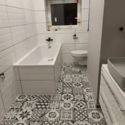 Wykończona łazienka z białą wanną obudowaną płytkami, toaletą, szarą umywalką nablatową i podłogą w czarno-białe wzorzyste płytki; na podłodze leży nóż do cięcia płytek.