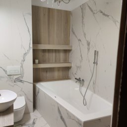 Kamil Kaszuba - Nowoczesna łazienka z wanną obudowaną płytkami imitującymi marmur, drewnianą wnęką z półkami i wiszącą lampą w stylu retro.