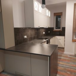 Kamil Kaszuba - Nowoczesna kuchnia z wyspą, białymi szafkami z podświetleniem, ciemnymi płytkami na ścianie i kolorową podłogą z paneli, oświetlona wiszącą lampą z okrągłą żarówką.