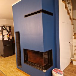 Kamil Kaszuba - Niebieski kominek z czarnym wkładem, z widocznym fragmentem salonu, podłogą z paneli i pojemnikiem z farbą obok, oświetlenie LED w suficie.