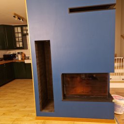 Kamil Kaszuba - Niebieska ściana działowa z wnękami na drewno i kominek, widoczna kuchnia w tle, świeżo położone panele podłogowe.