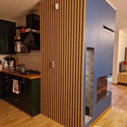 Kamil Kaszuba - Nowoczesna kuchnia z drewnianą ścianką działową z pionowych listew i zabudowanym kominkiem w kolorze granatowym, widoczny fragment podłogi z paneli drewnianych.