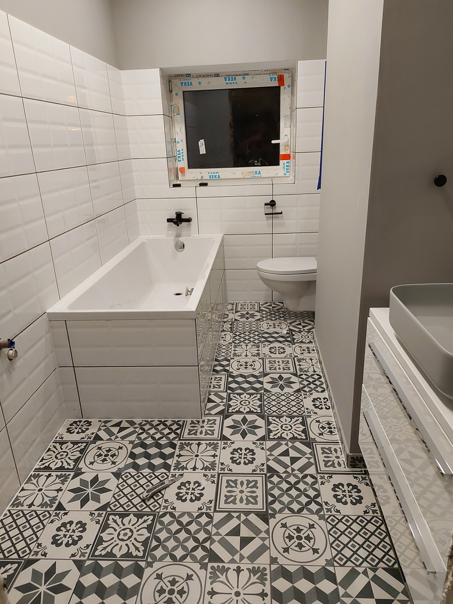 Wykończona łazienka z białą wanną obudowaną płytkami, toaletą, szarą umywalką nablatową i podłogą w czarno-białe wzorzyste płytki; na podłodze leży nóż do cięcia płytek.