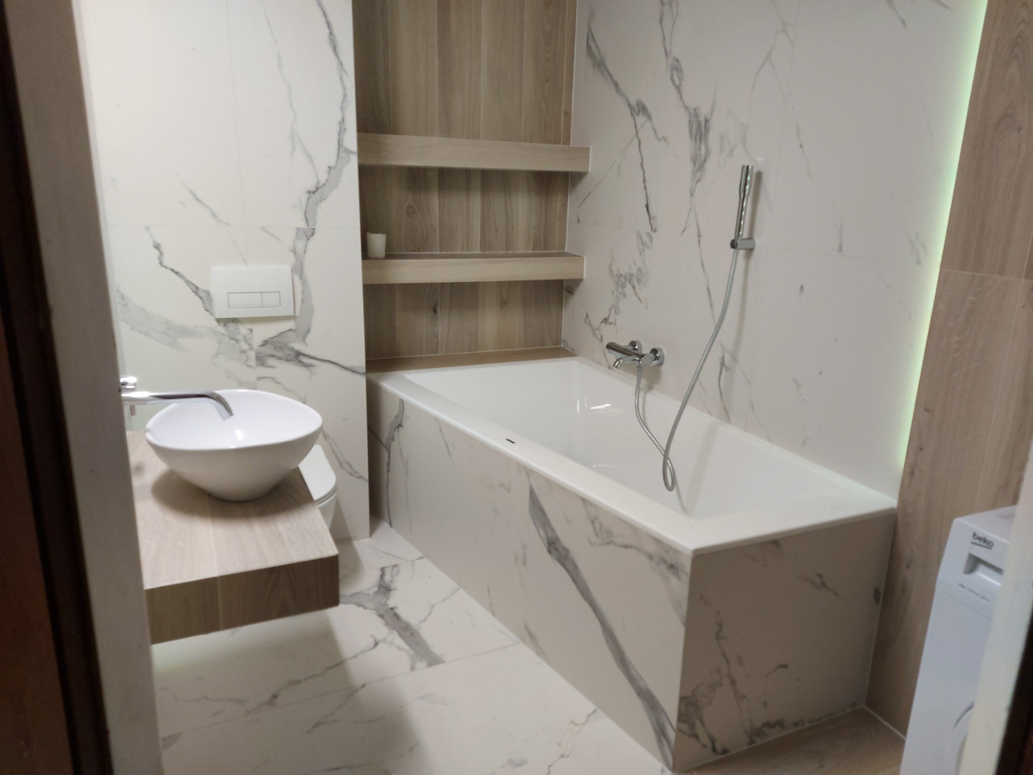 Nowoczesna łazienka z wanną obudowaną marmurowymi płytami, umywalką nablatową na drewnopodobnym blacie i półkami w zabudowie. Ściany wykończone płytkami imitującymi marmur i drewno.