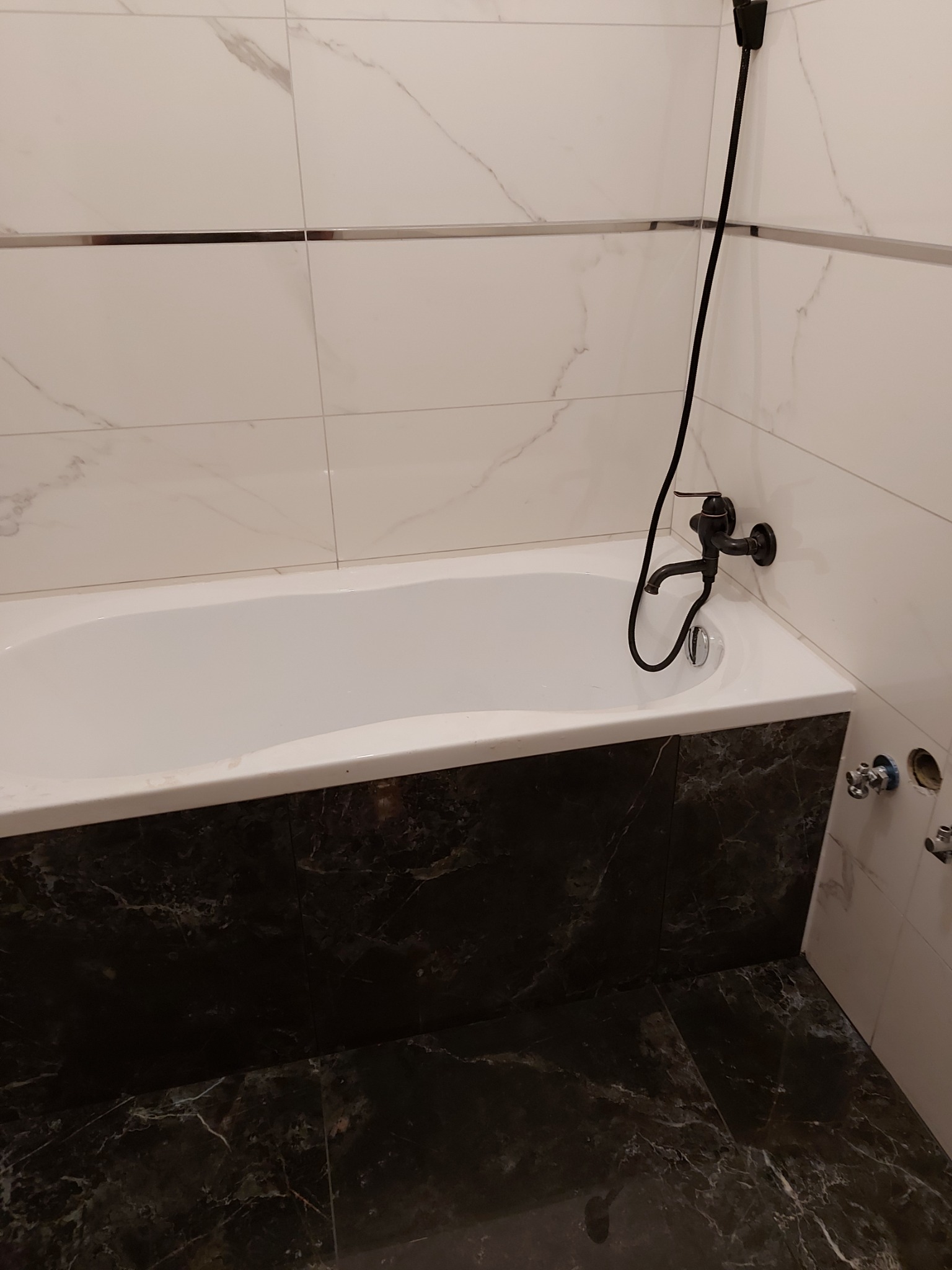 Biała wanna z czarną armaturą i obudową z płytek imitujących czarny marmur, ściany wyłożone białymi płytkami z delikatnym wzorem marmuru i metalowymi listwami dekoracyjnymi.