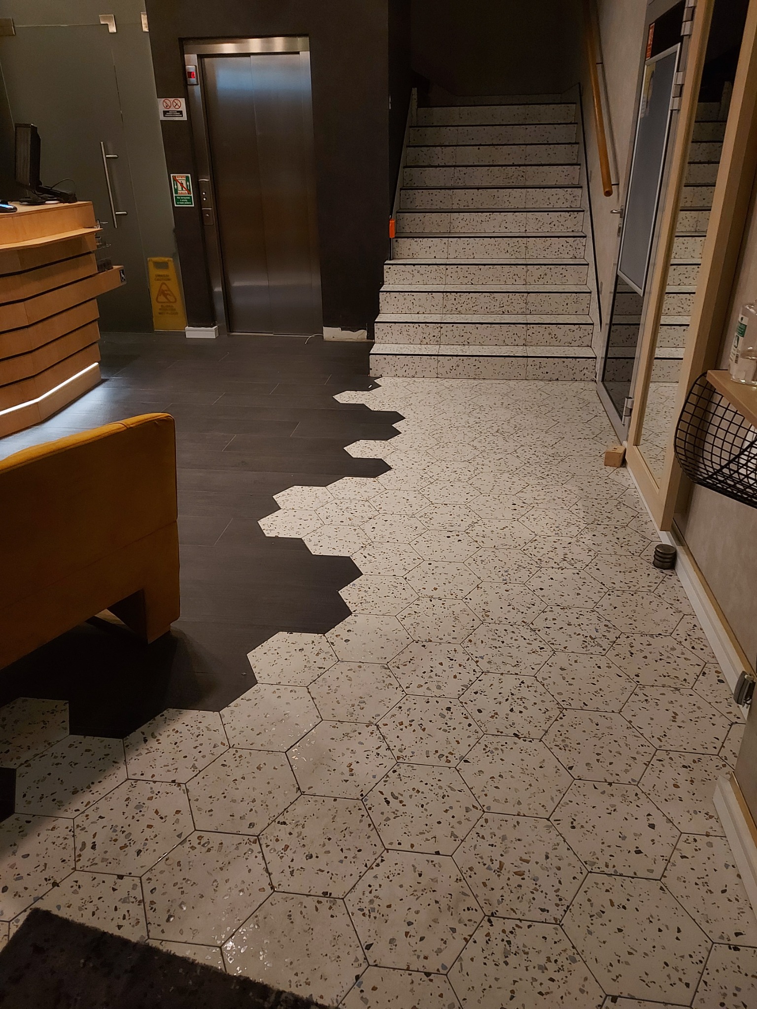 Kontrastowe ułożenie podłogi: heksagonalne płytki lastryko przechodzą w ciemne panele, widoczny fragment sofy, schody i winda w tle.