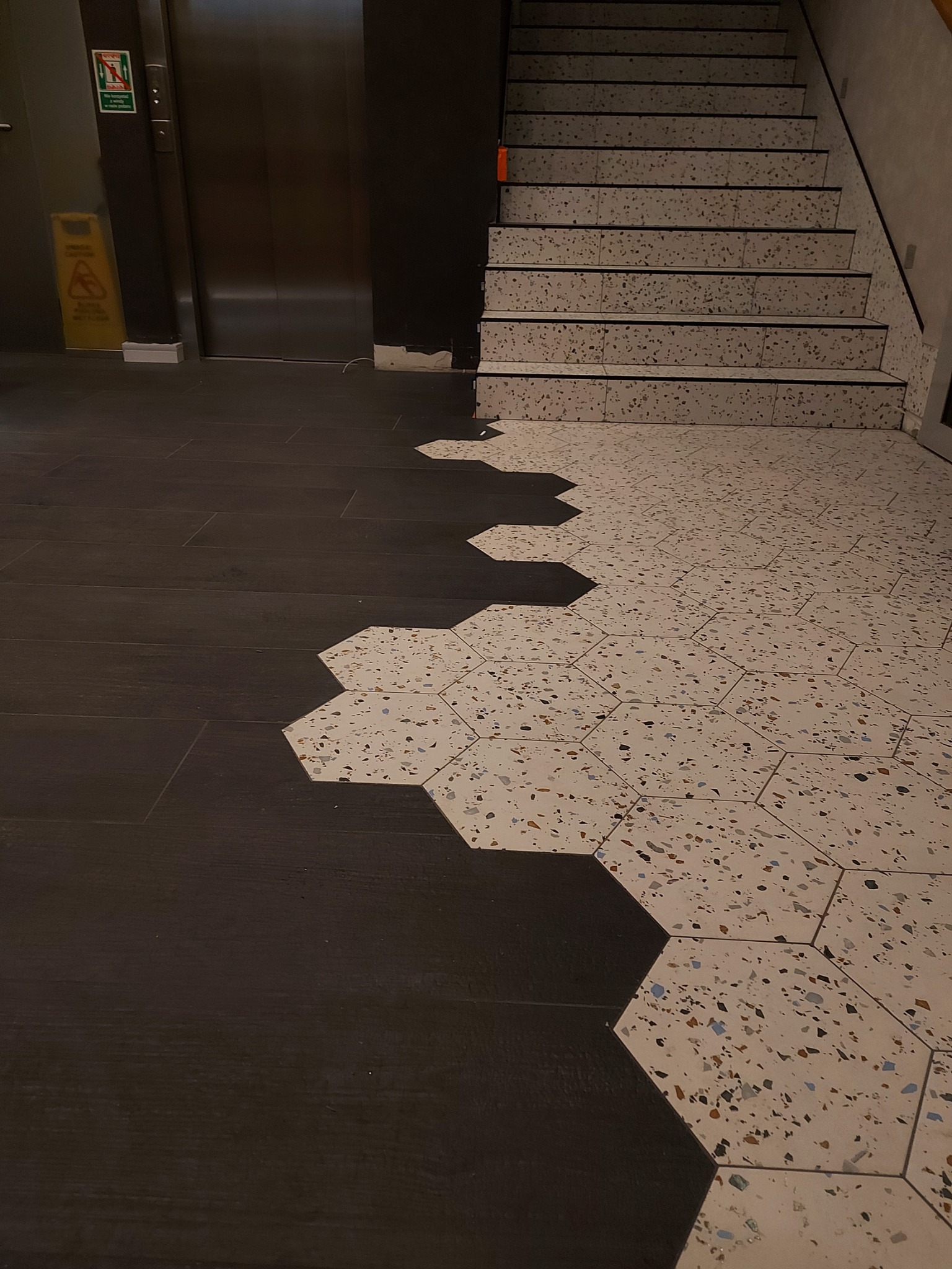 Połączenie ciemnych paneli podłogowych i jasnych, heksagonalnych płytek lastryko na podłodze, prowadzące do schodów z lastryko i windy.