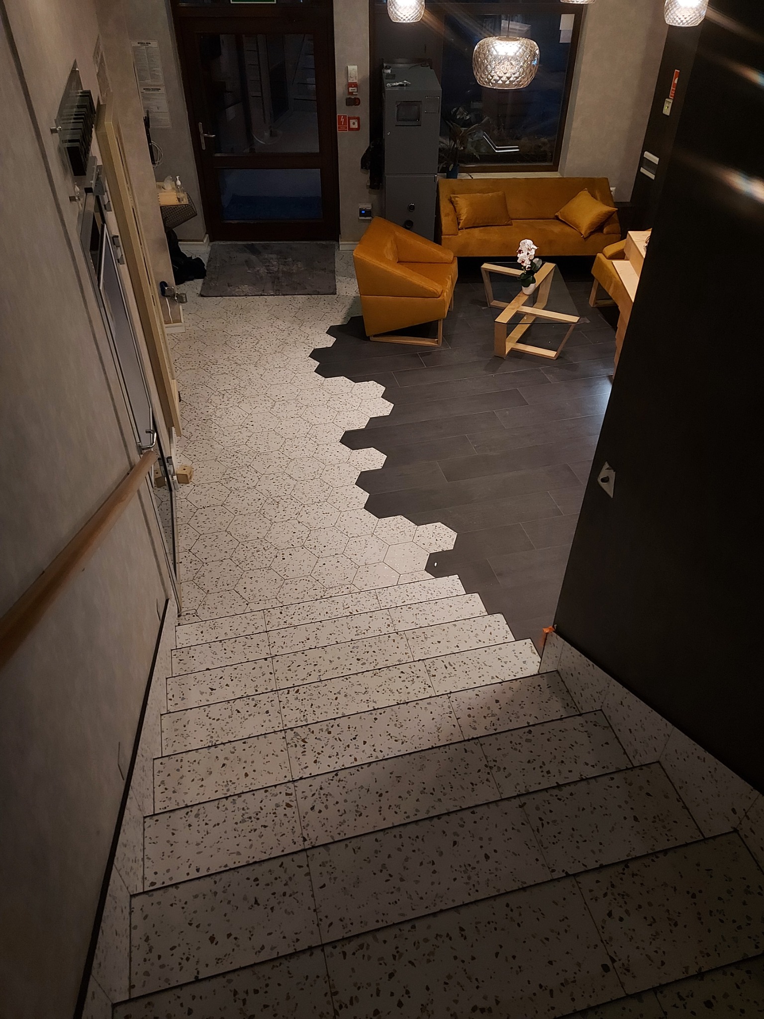 Widok z góry na schody wyłożone płytkami lastryko przechodzące w podłogę z ciemnych paneli, z fragmentem salonu z sofą i fotelem w odcieniach musztardowych w tle.