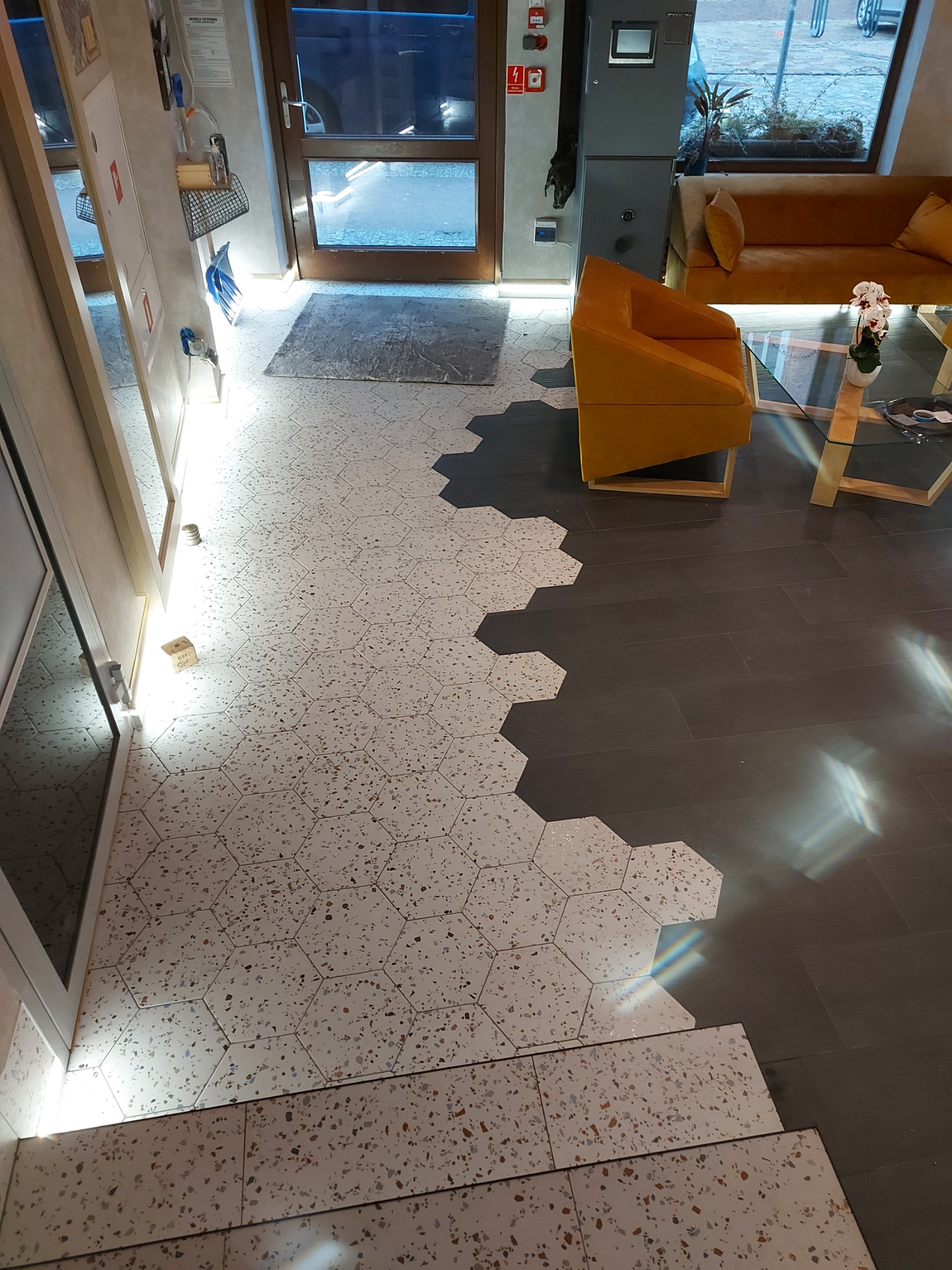 Wnętrze z heksagonalną mozaiką podłogową przechodzącą w ciemne panele, widoczny fragment sofy i fotela w odcieniach musztardowych oraz szklany stolik kawowy.