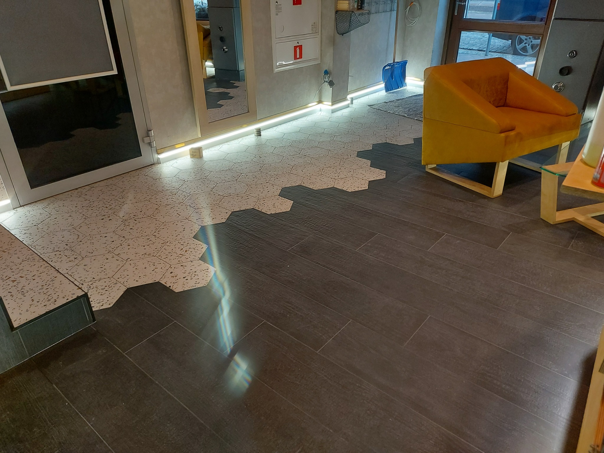 Połączenie heksagonalnych płytek podłogowych w kolorze terazzo z ciemnymi panelami podłogowymi imitującymi drewno, oświetlenie LED przy ścianie, żółty fotel w tle.