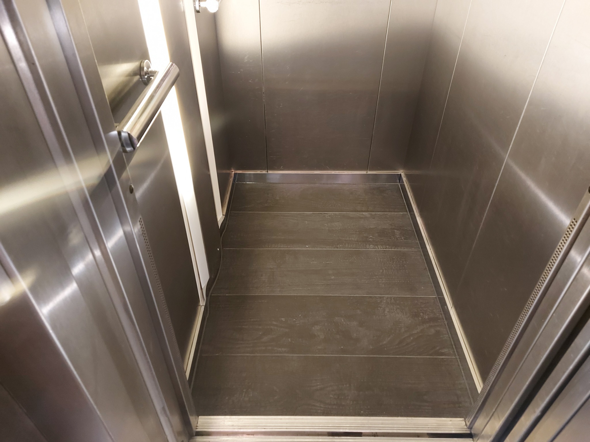 Wnętrze kabiny prysznicowej z wyłożonymi ciemnymi płytkami podłogowymi i ścianami ze stali nierdzewnej, widoczny uchwyt i oświetlenie.