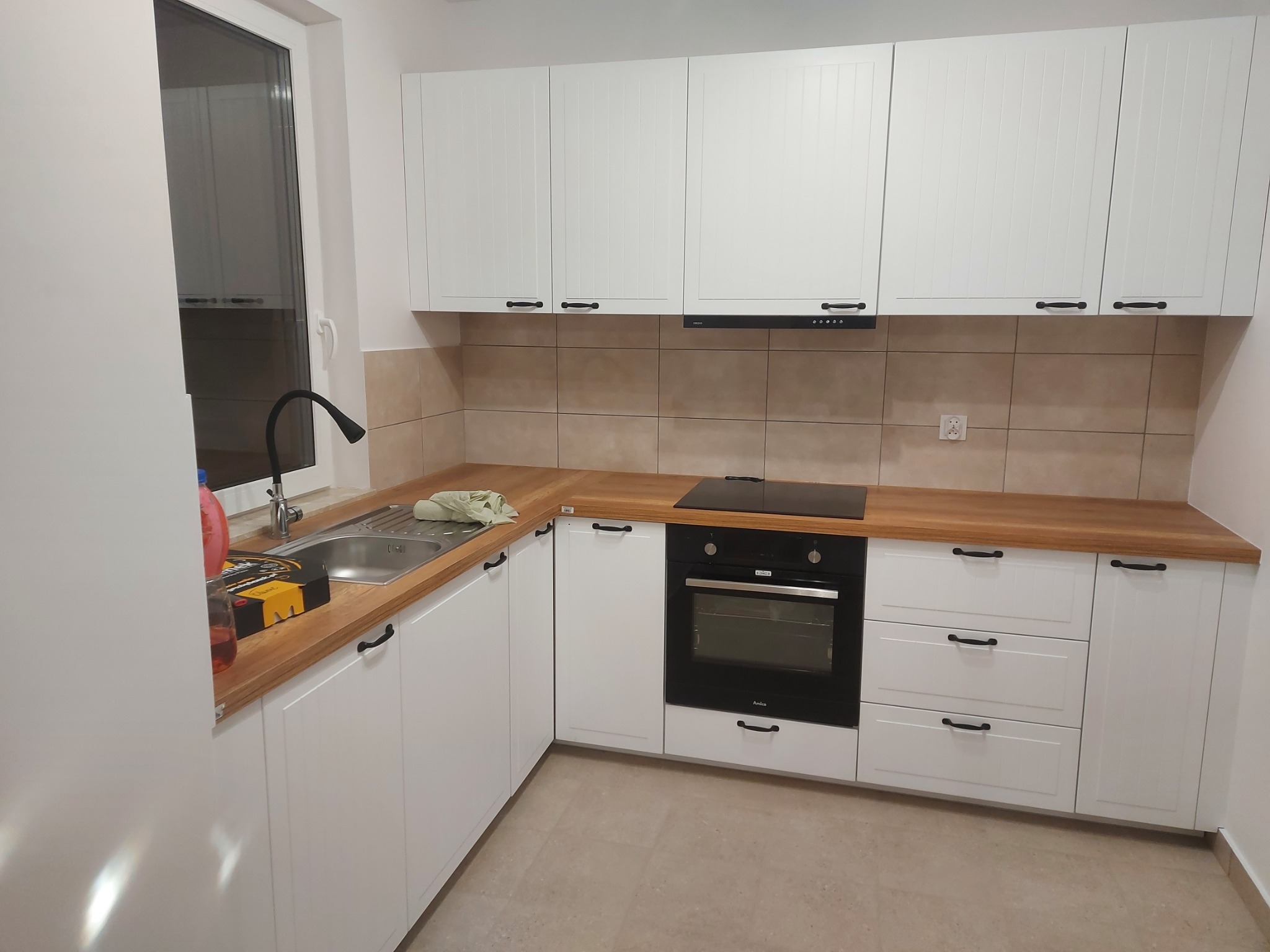 Nowoczesna, jasna kuchnia z białymi szafkami z frezowanymi frontami i drewnianym blatem. Na blacie przy zlewozmywaku leżą narzędzia i ściereczka. Widoczny piekarnik i płyta grzewcza.