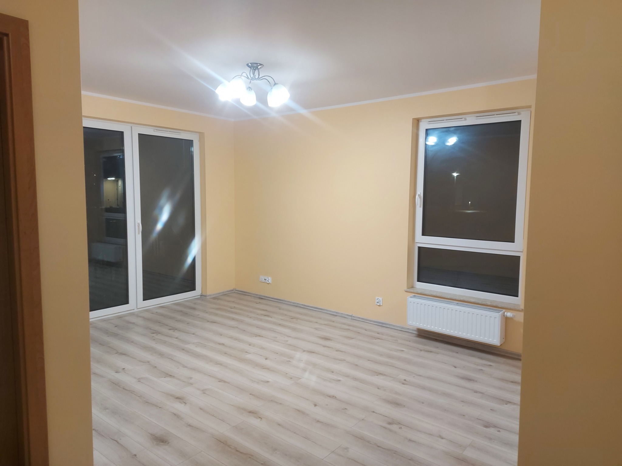 Pusty pokój z panelami podłogowymi w odcieniach szarości, świeżo pomalowane ściany w odcieniach beżu, białe okno z grzejnikiem pod spodem i przesuwne drzwi balkonowe.