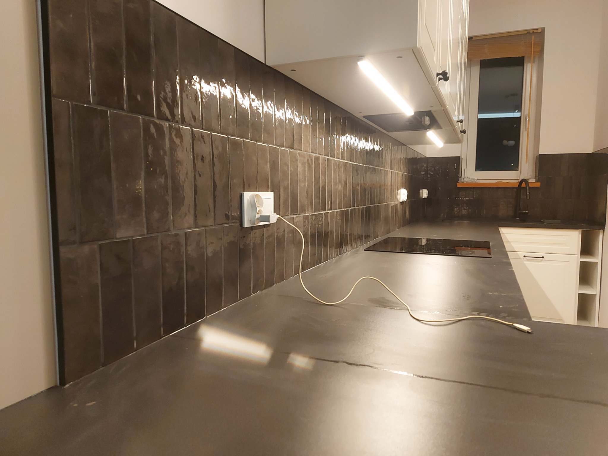 Ciemnobrązowe, błyszczące płytki ułożone pionowo na ścianie w kuchni, widoczny fragment blatu, gniazdko elektryczne z podłączoną ładowarką, białe szafki kuchenne z oświetleniem LED podszafkowym.