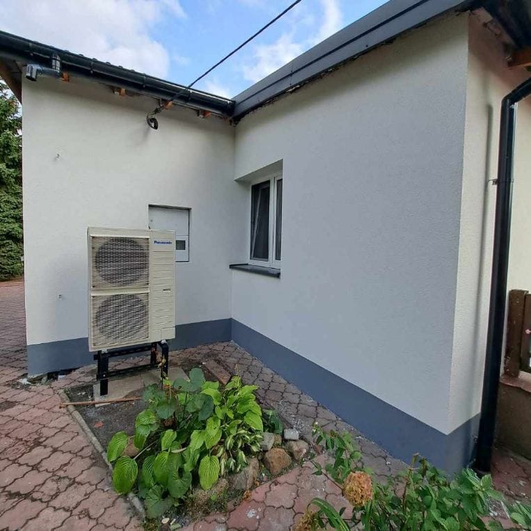 Biała elewacja domu z szarą podmurówką i zamontowaną pompą ciepła Panasonic, widok z boku na tle kostki brukowej i roślinności.