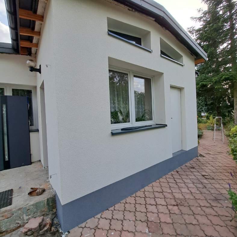 Biały budynek z nową elewacją i oknami, widoczne świeże tynki, szara podmurówka i chodnik z kostki brukowej. Na schodach leżą buty, a w oknie widać rośliny.