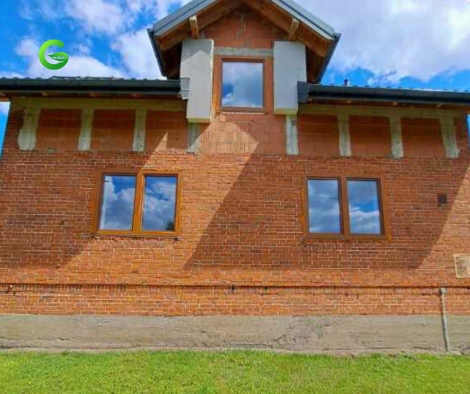 Surowy stan elewacji budynku z czerwonej cegły z zamontowanymi oknami drewnianymi, widoczne elementy konstrukcyjne i przygotowanie do dalszych prac wykończeniowych.