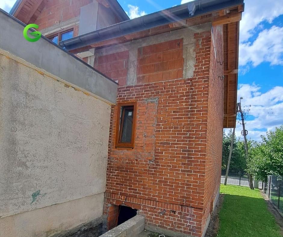 Częściowo otynkowany budynek z cegły, z widocznym oknem z brązową ramą, fragment elewacji w trakcie prac wykończeniowych, widoczne pustaki i fragment ocieplenia, błękitne niebo w tle.