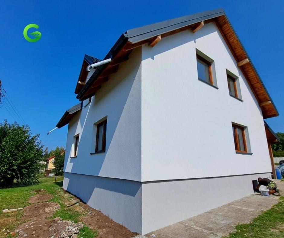 Świeżo otynkowany dom jednorodzinny z białymi ścianami i szarą podmurówką, widoczne drewniane elementy pod dachem, okna z brązowymi ramami i pracownik wykonujący prace wykończeniowe przy fundamencie.