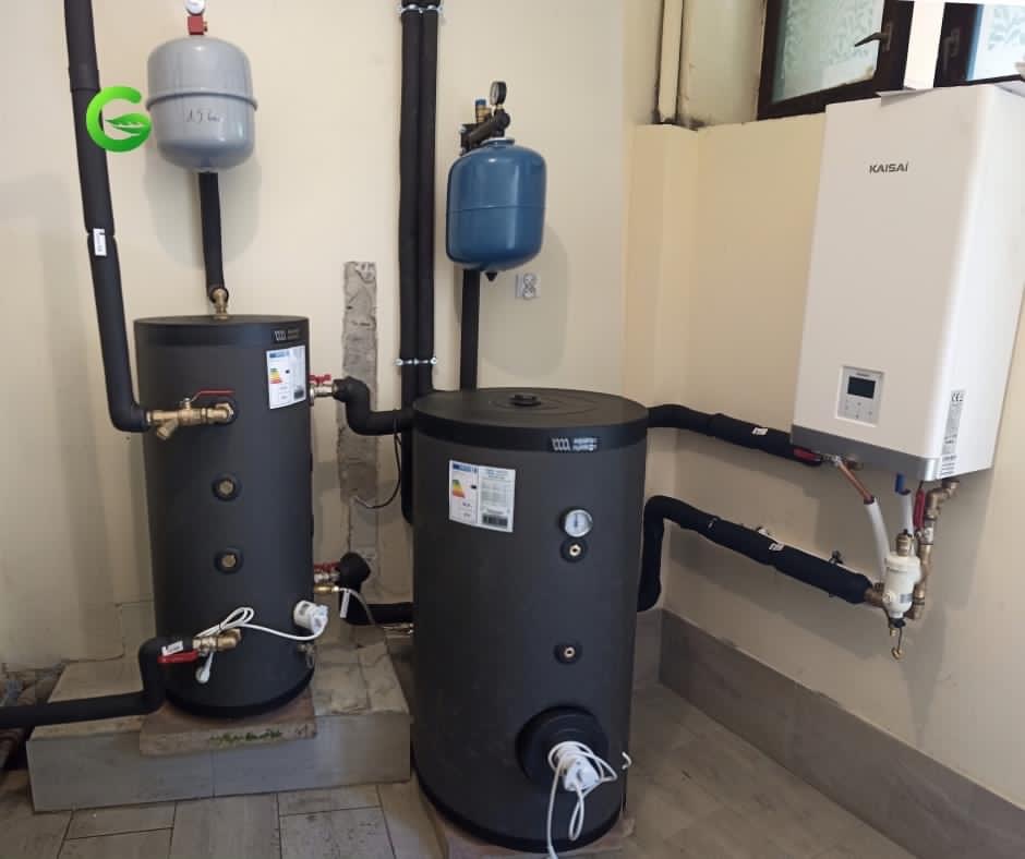 Dwa ciemnoszare zbiorniki akumulacyjne ciepłej wody, z podłączonymi rurami w izolacji, obok białego modułu pompy ciepła Kaisai zamontowanego na ścianie w jasnym pomieszczeniu.