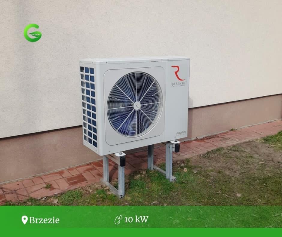 Zewnętrzna jednostka pompy ciepła Rotenso Aquami 10kW zamontowana na metalowych wspornikach przy elewacji budynku z widocznym logo firmy.