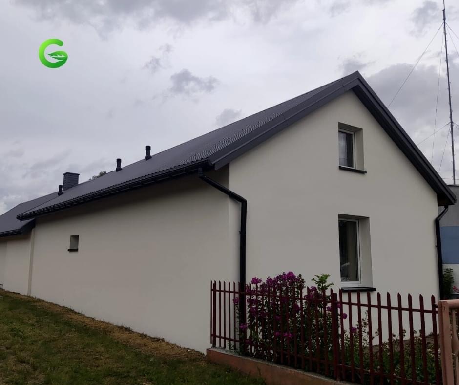 Jasna elewacja domu jednorodzinnego z ciemnym dachem i orynnowaniem, widok z niskiej perspektywy, z fragmentem czerwonego ogrodzenia i kwiatami na pierwszym planie, logo firmy w lewym górnym rogu.