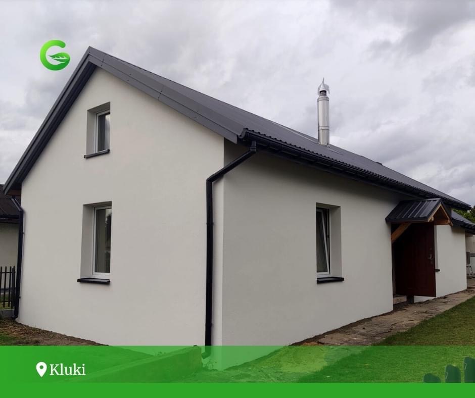 Dom jednorodzinny po remoncie elewacji, z nową, jasną, gładką powłoką, czarnym dachem i rynnami, widoczny komin ze stali nierdzewnej, pochmurne niebo w tle.