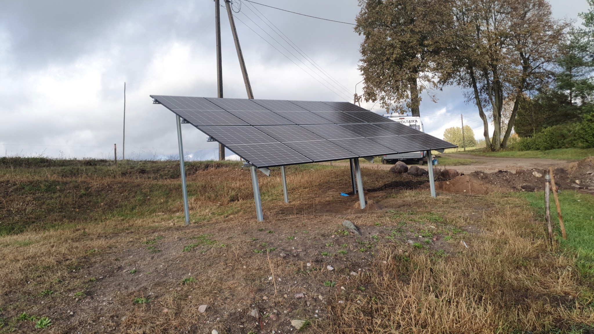 Panel słoneczny ustawiony na metalowych podporach na nierównym terenie z trawą i ziemią, słup elektryczny w tle, pochmurne niebo.