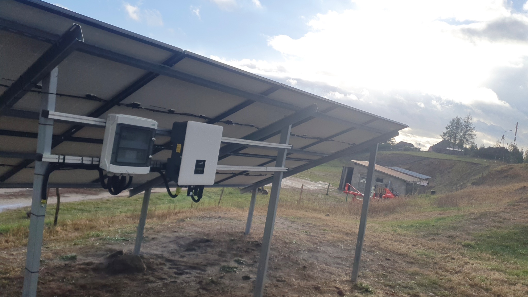 Widok od dołu na panele słoneczne z zamontowanymi inwerterami, na tle wiejskiego krajobrazu z budynkami gospodarczymi i pochmurnym niebem.