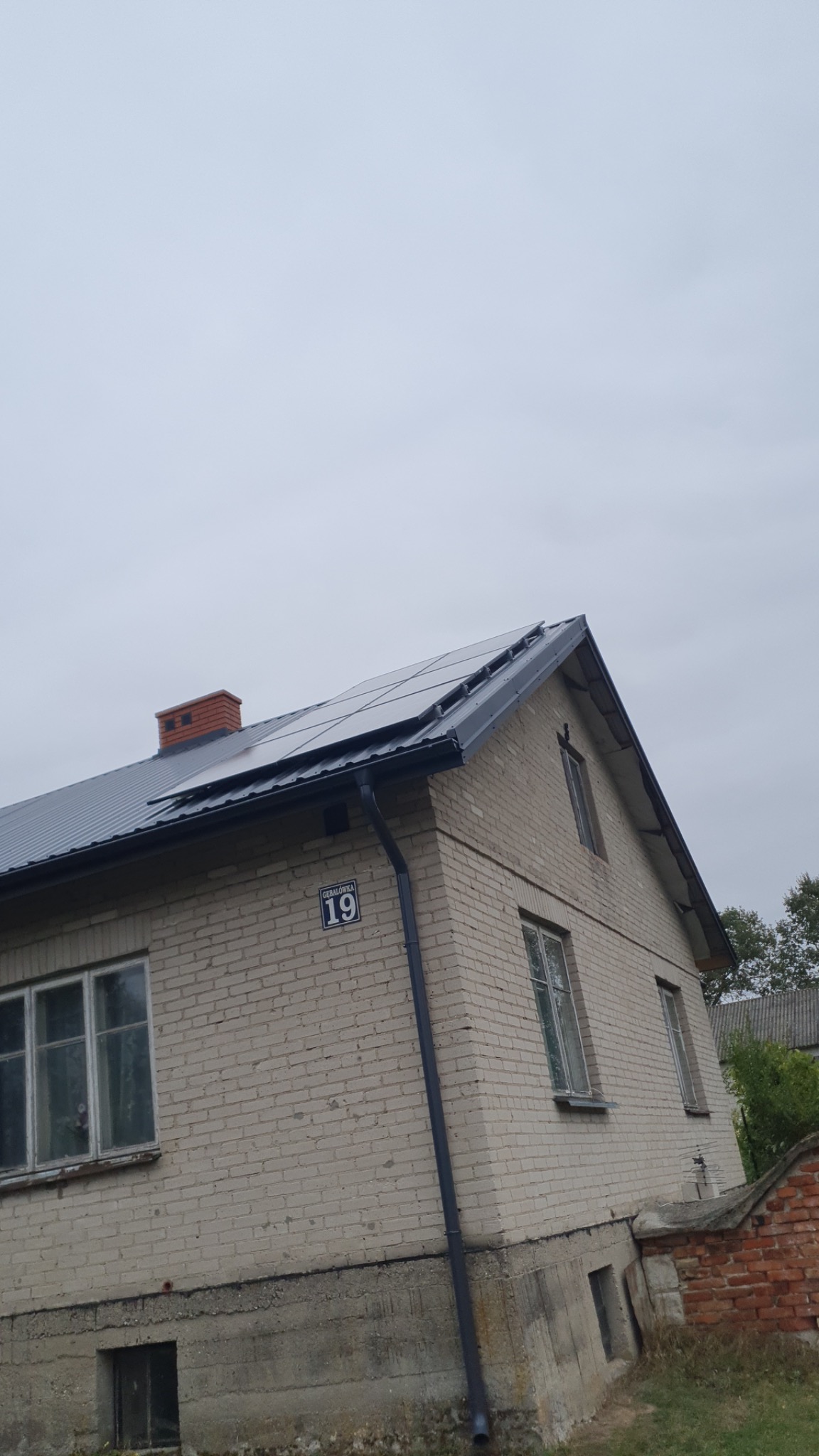 Dom z jasnej cegły z nowymi panelami na dachu i numerem 19 na ścianie, widziany z dołu pod pochmurnym niebem.