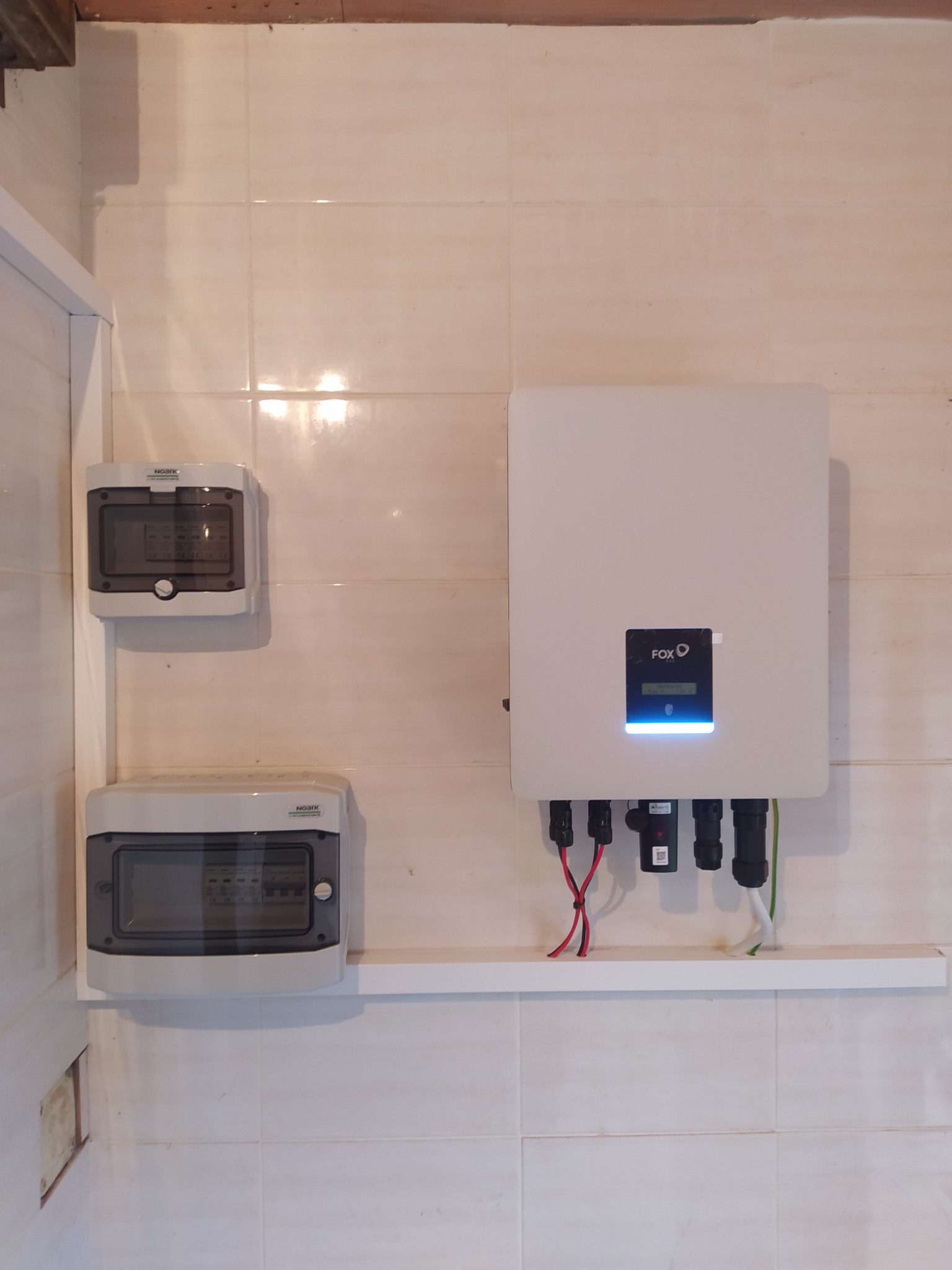 Biały falownik FOX z podłączonymi przewodami i dwoma rozdzielnicami Noark zamontowany na kafelkowanej ścianie.