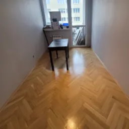 Pokój po remoncie z parkietem ułożonym w jodełkę, szare ściany, widok na okno z parapetem zastawionym przedmiotami, czarny stolik przy ścianie.