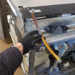 Serwisant w Krakowie w czarnych rękawicach w kropki demontuje wężyk wewnątrz otwartej zmywarki, widoczne elementy wewnętrzne urządzenia i inne przewody.