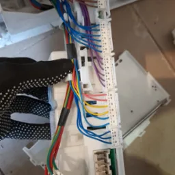 Moduł sterujący pralki z podłączonymi wiązkami kabli w r&oacute;żnych kolorach, trzymany w dłoni w roboczej rękawicy w kropki.