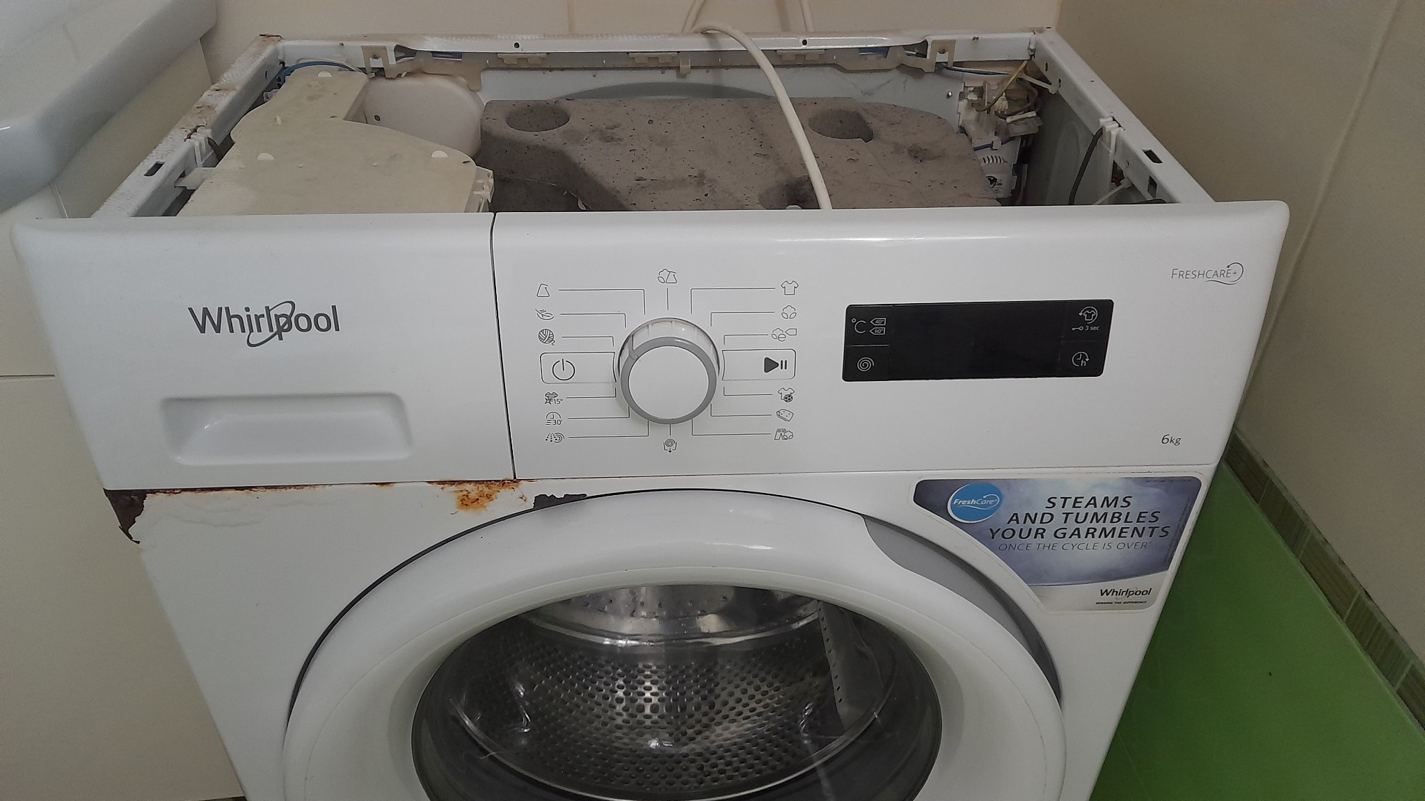 Uszkodzona pralka Whirlpool z otwartą obudową, widoczne ślady rdzy i zniszczeń, wnętrze brudne, ukazujące stan przed naprawą.
