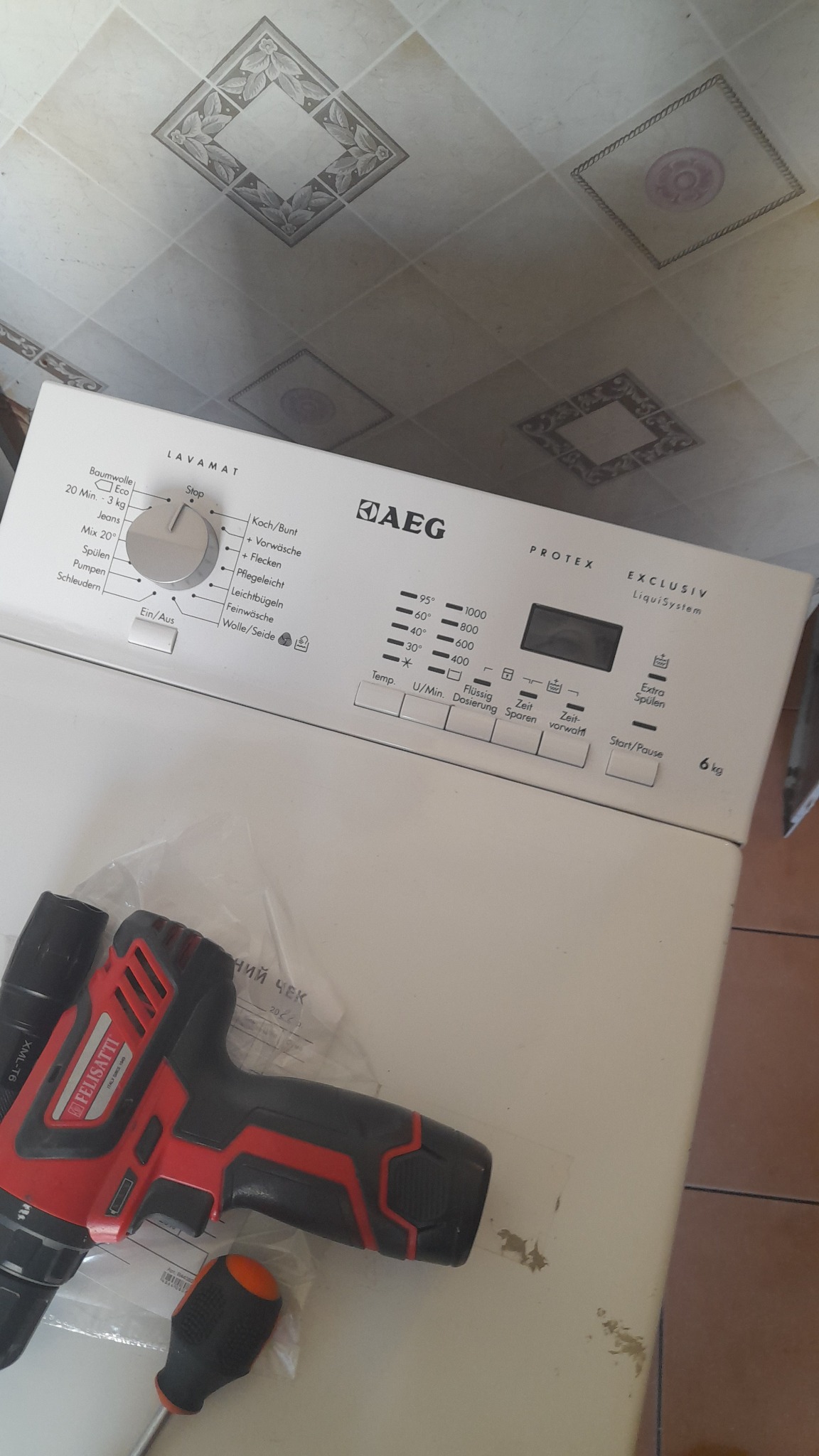 Pralka marki AEG z widocznym panelem sterowania, obok leży wkrętarka akumulatorowa Felisatti i śrubokręt z pomarańczową rączką, tło stanowią kafelki na ścianie i podłodze.
