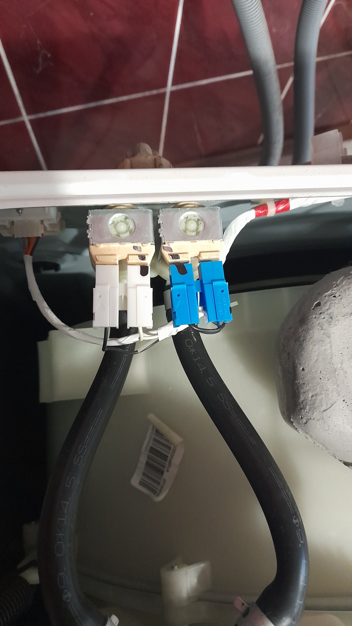 Wnętrze pralki z widocznymi zaworami elektromagnetycznymi, przewodami doprowadzającymi wodę i instalacją elektryczną; zawory z niebieskimi i białymi złączkami.