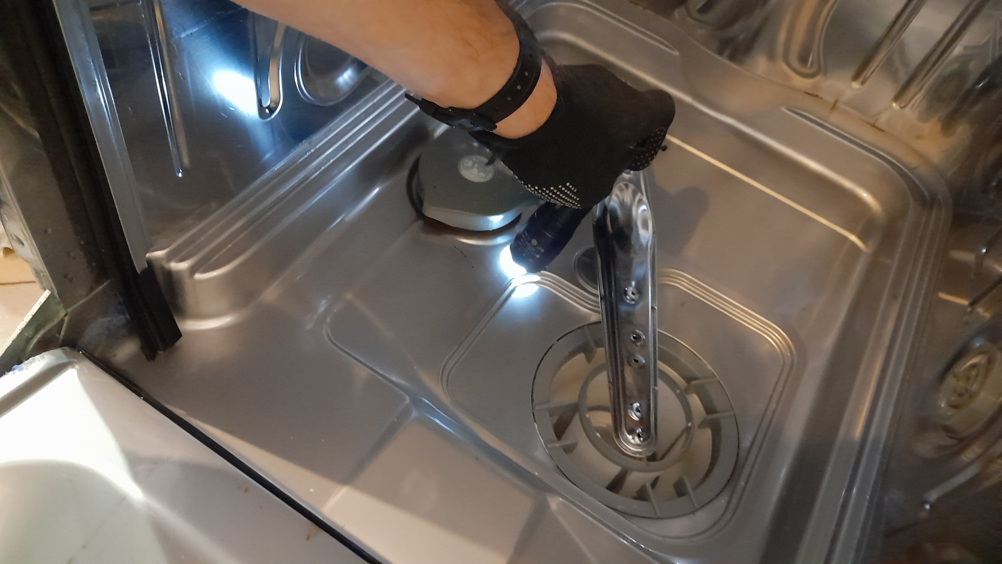 Wnętrze zmywarki z widocznym ramieniem natryskowym i filtrem, oświetlone punktowo przez osobę w czarnej rękawicy podczas inspekcji.
