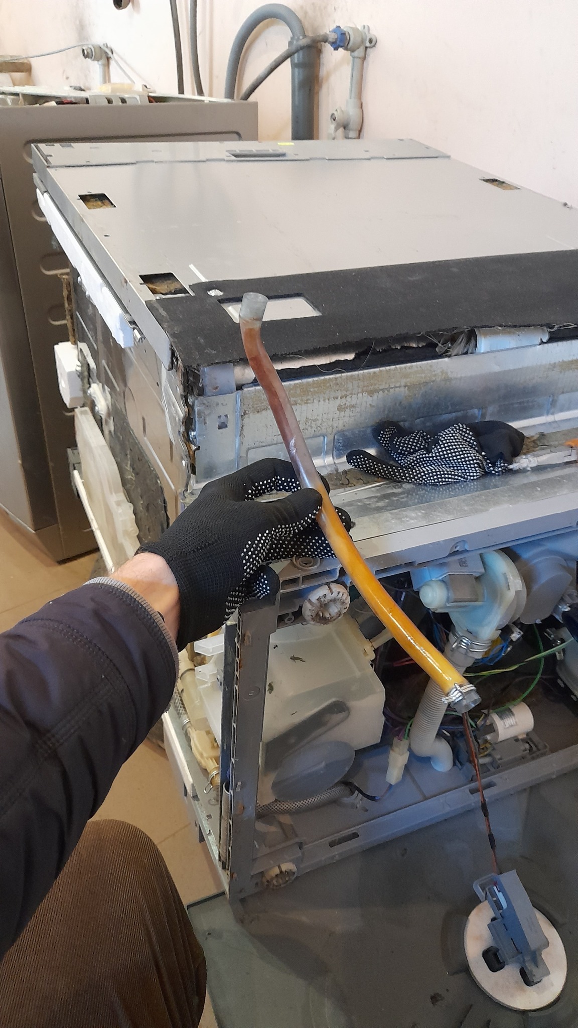 Serwisant w Krakowie w czarnych rękawicach w kropki demontuje wężyk wewnątrz otwartej zmywarki, widoczne elementy wewnętrzne urządzenia i inne przewody.