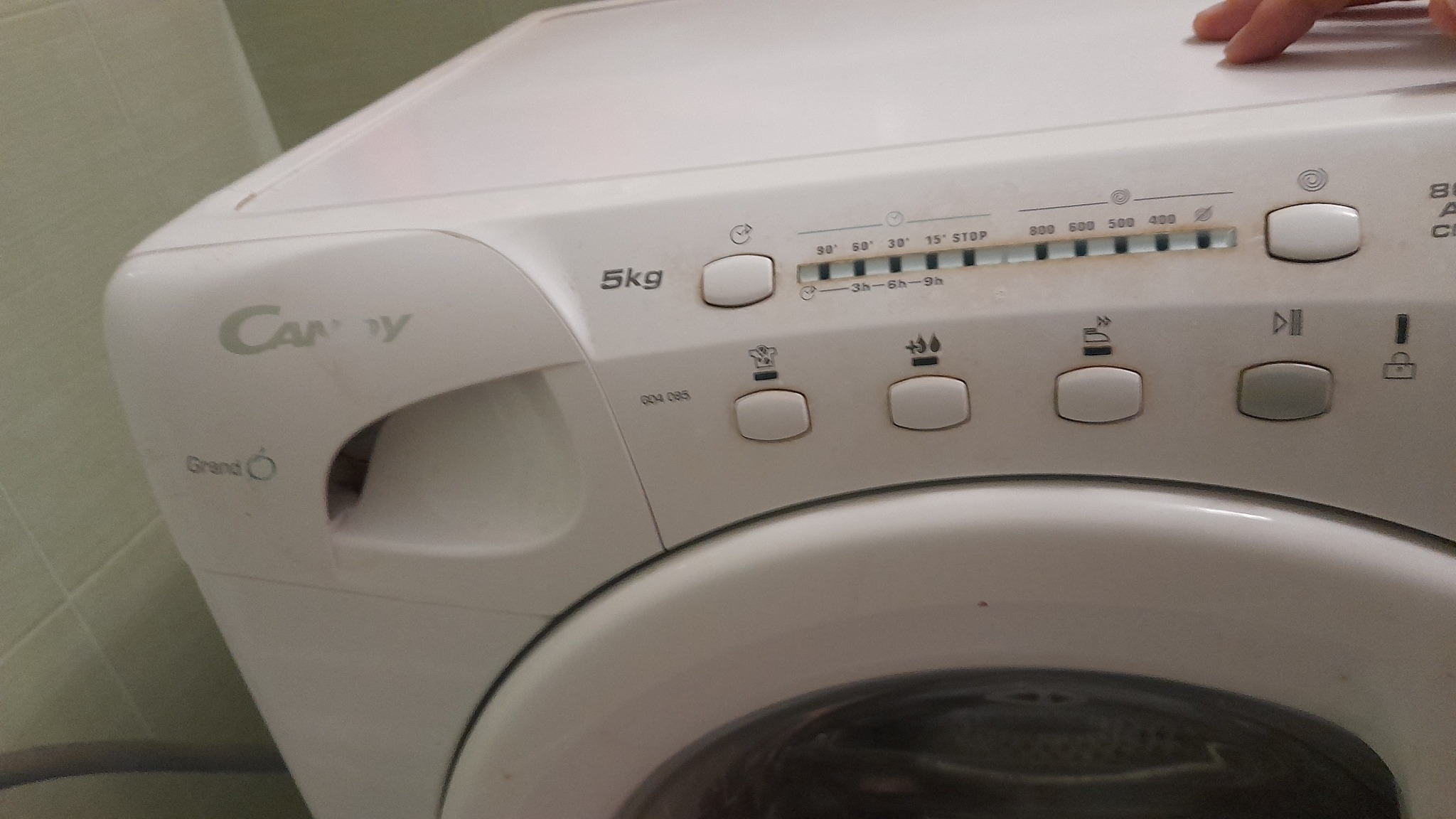 Panel sterowania pralki Candy Grand O z widocznymi przyciskami, pokrętłem wyboru temperatury i programami prania, z delikatnym ujęciem palca na górnej powierzchni.