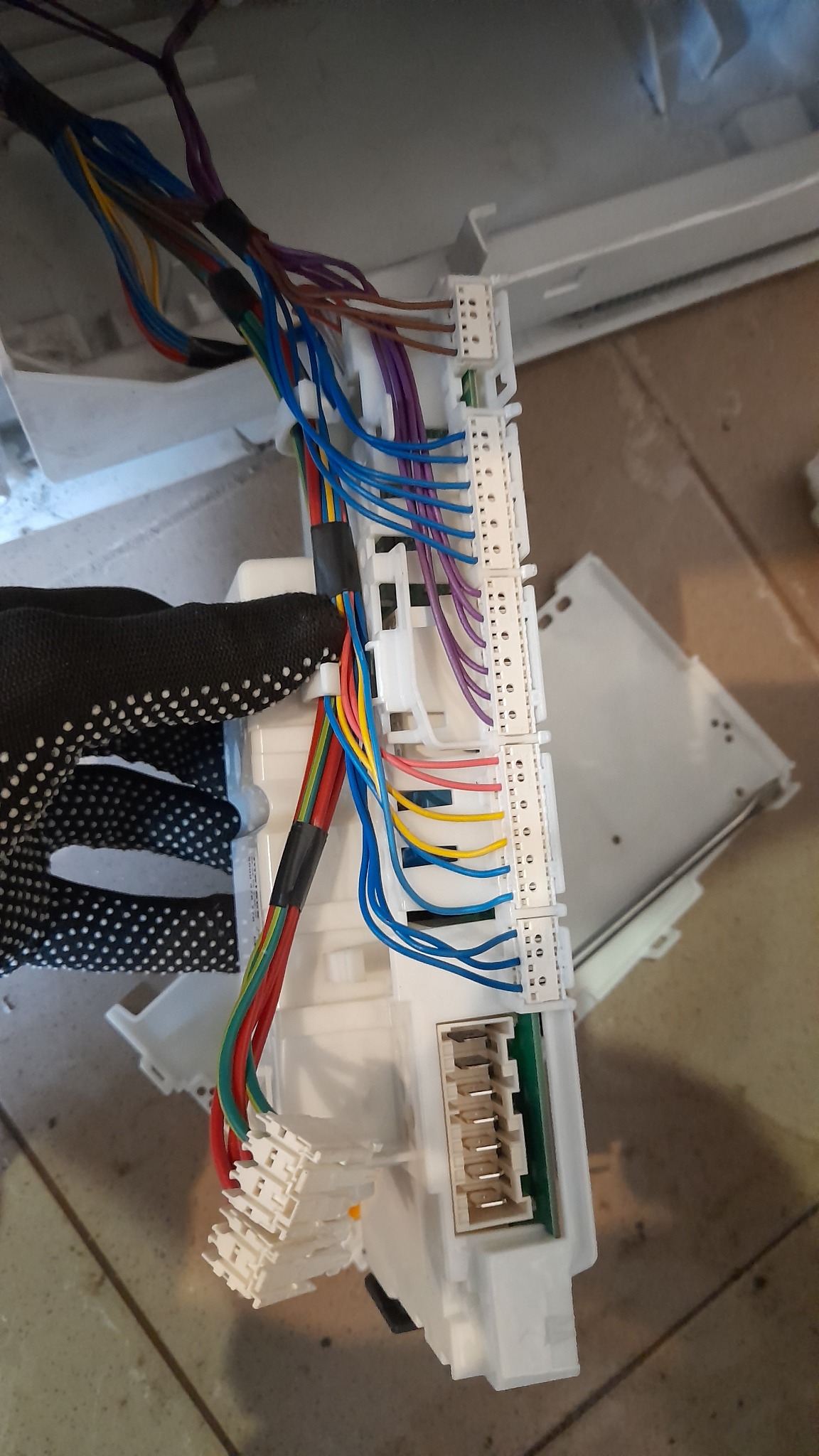 Moduł sterujący pralki z podłączonymi wiązkami kabli w różnych kolorach, trzymany w dłoni w roboczej rękawicy w kropki.