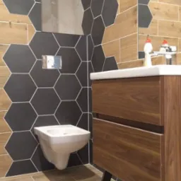 Nowoczesna łazienka z heksagonalnymi płytkami w odcieniach szarości i drewna, wisząca toaleta i umywalka z szafką z ciemnego drewna.
