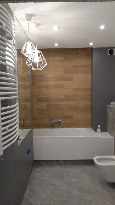 Nowoczesna łazienka z drewnopodobnymi płytkami na ścianie, białą wanną, grzejnikiem drabinkowym i geometrycznymi lampami wiszącymi.