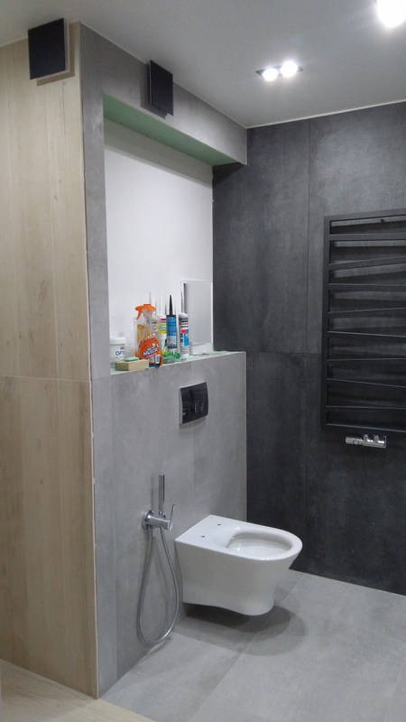 Nowoczesna łazienka z wiszącą toaletą, bidetką, grzejnikiem drabinkowym i szarą armaturą, widoczne materiały budowlane na półce w zabudowie.