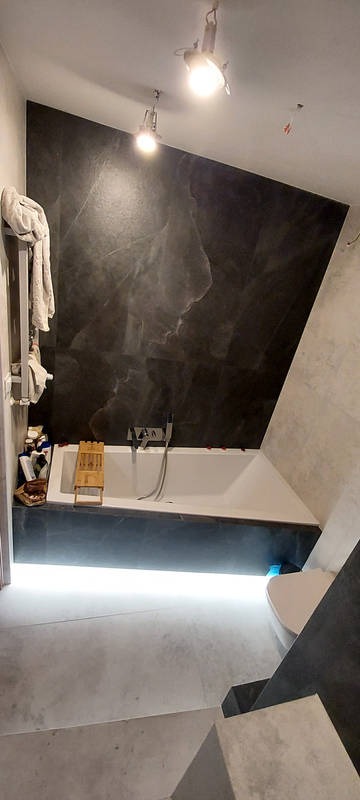Nowoczesna łazienka z asymetryczną ścianą pokrytą czarnym marmurem, podświetlana wanna z drewnianą półką, minimalistyczne oświetlenie punktowe i betonowa podłoga.
