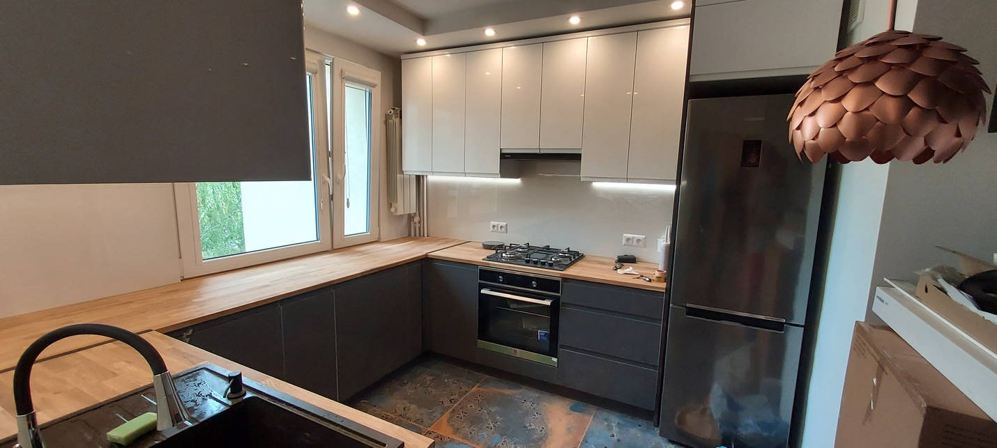 Nowoczesna kuchnia w szaro-białej kolorystyce z drewnianym blatem, widok na okno z roletą, ozdobna lampa w kształcie szyszki, ciemna podłoga z nieregularnym wzorem.