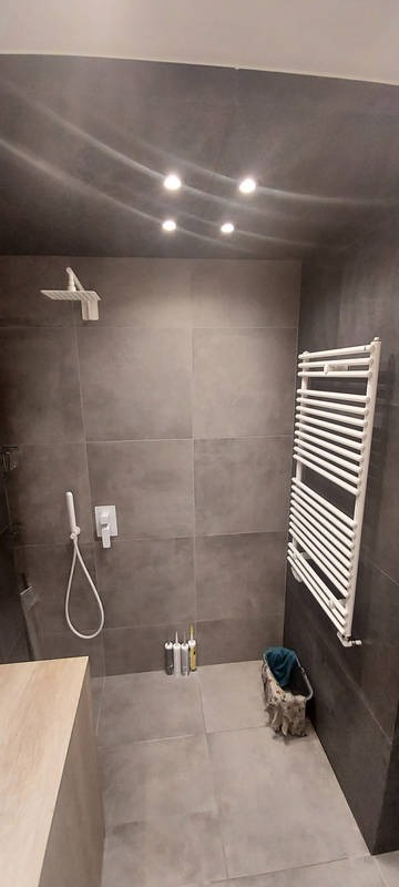 Nowoczesna łazienka z szarymi płytkami, deszczownicą, grzejnikiem drabinkowym i tubami z silikonem na podłodze, oświetlona czterema punktowymi lampami w suficie.