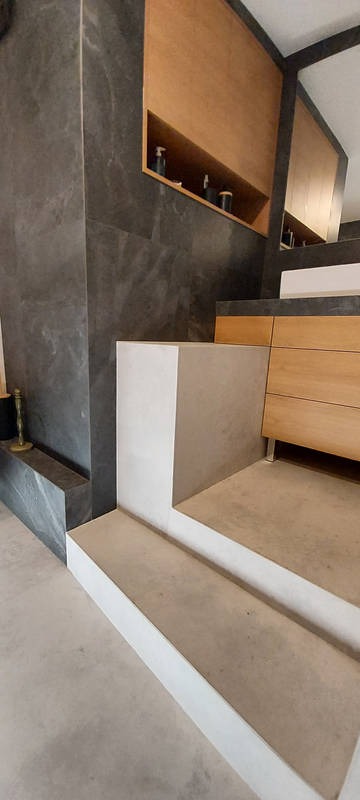 Nowoczesna łazienka z szarą, marmurkową ścianą, drewnianymi szafkami i półkami, oraz betonowymi schodkami prowadzącymi do wyższego poziomu, z widocznym złotym wazonem.