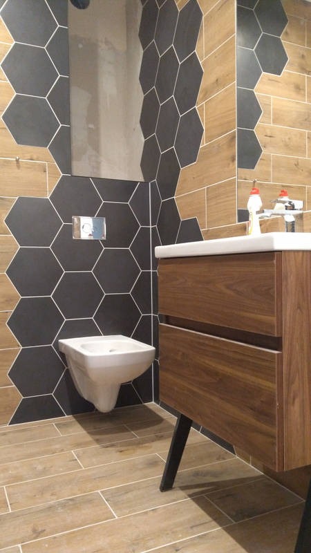Nowoczesna łazienka z heksagonalnymi płytkami w odcieniach szarości i drewna, wisząca toaleta i umywalka z szafką z ciemnego drewna.